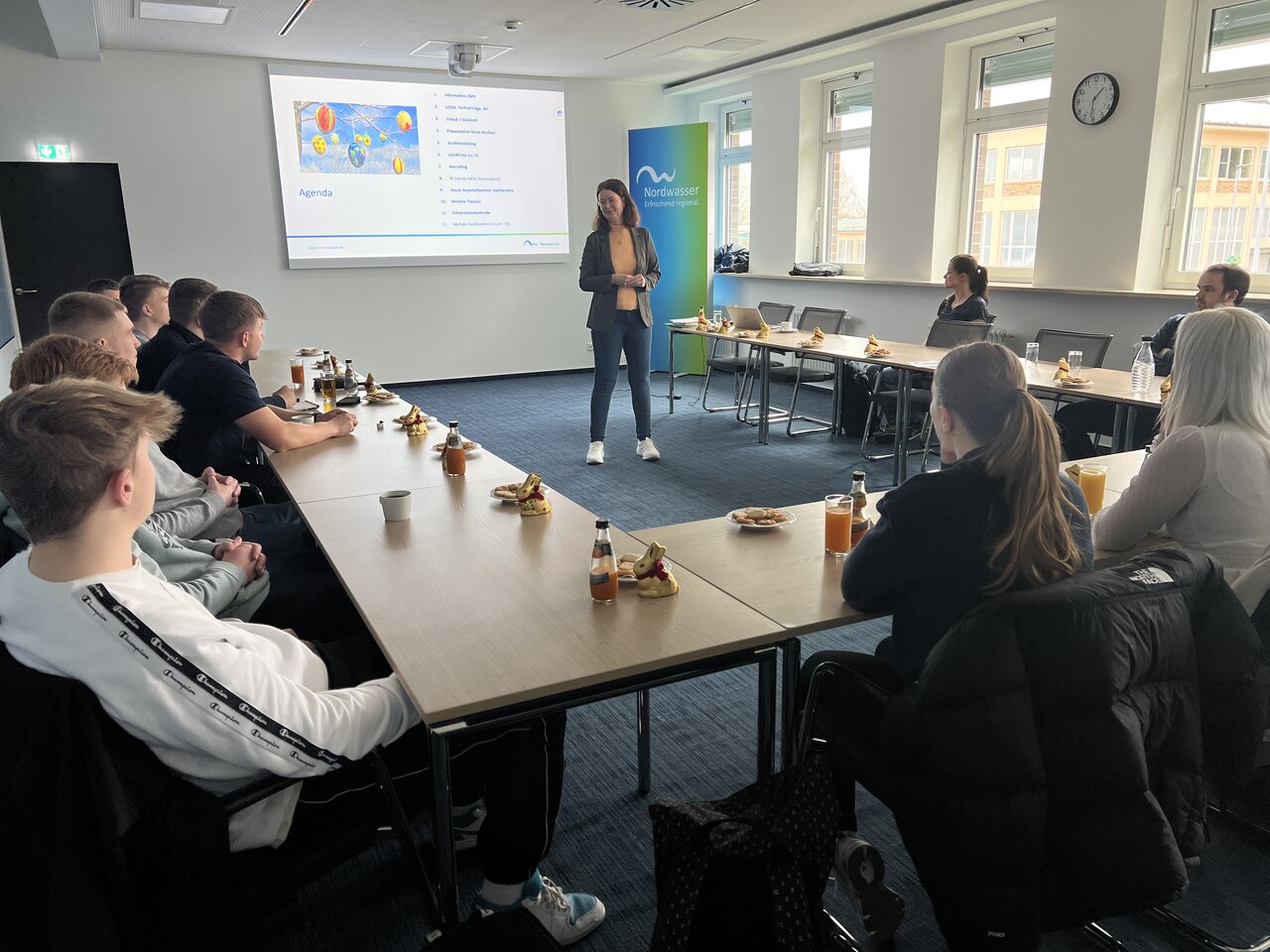 Eine Gruppe von Personen, die an einer U-förmigen Tafel sitzen und sich eine Präsentation anschauen. 