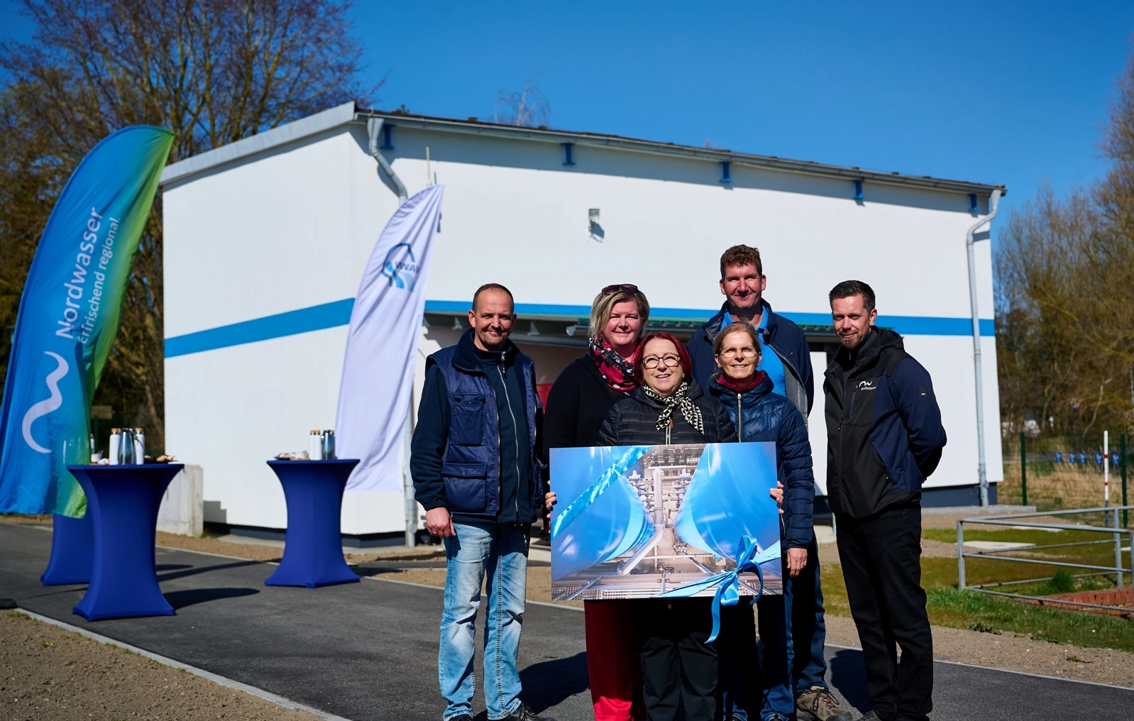 Drei Frauen und drei Männer stehen vor dem Wasserwerksgebäude welches weiß ist und halten ein großes Bild in die Kamera, welches das innere des Wasserwerkes zeigt