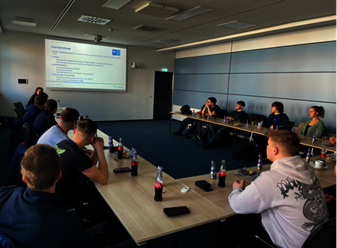 Azubis sitzen an einer Tafel in U-Form uns schauen auf einen Bildschirm an der Wand.