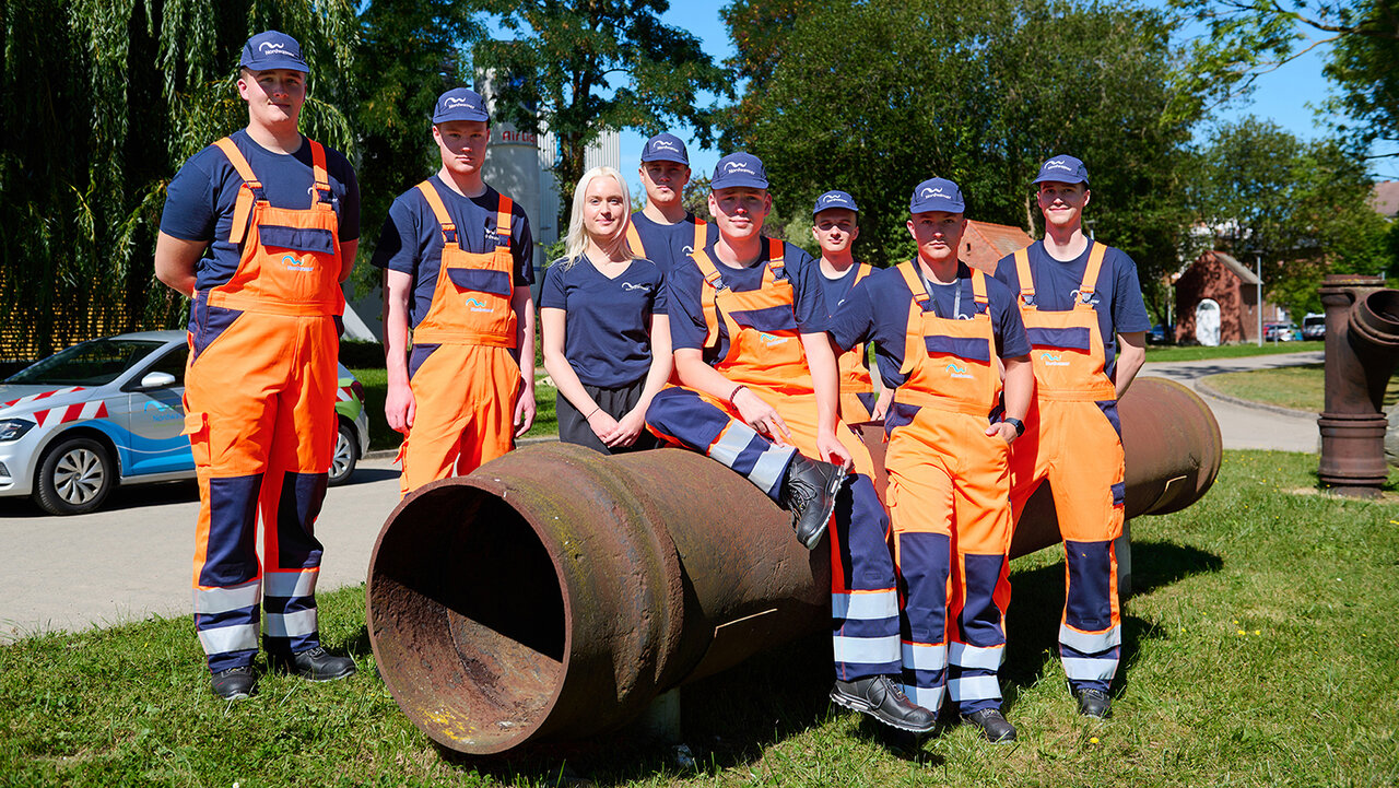 Gruppenfoto von vielen Jungen Leuten, die in Arbeitsbekleidung vor und auf einem großen Rohr posieren.